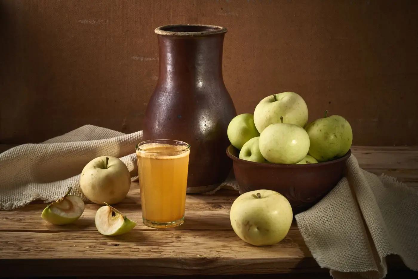 Glass of apple juice beside green apples and a clay jug on wood