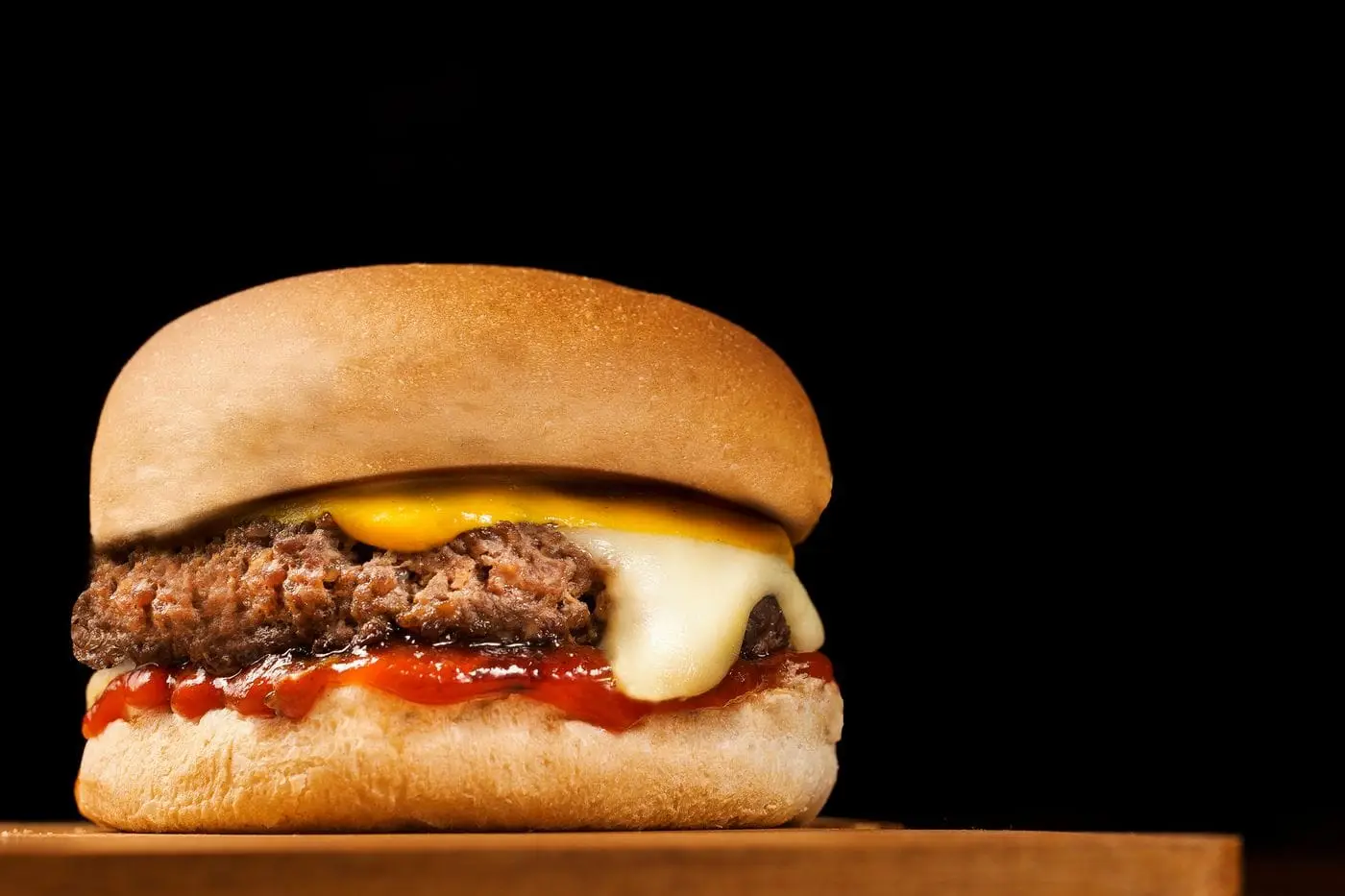 Cheeseburger with melted cheese and ketchup on a wooden board against black background