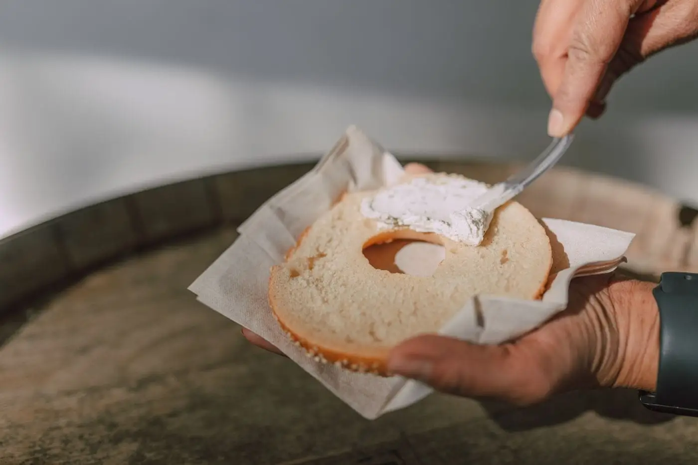 Hands spreading cream cheese on a sliced bagel half with a knife