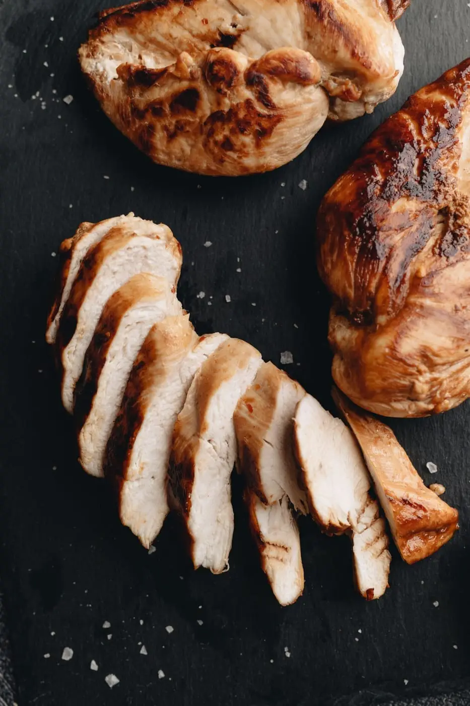 Sliced and whole grilled chicken breasts on a dark slate board
