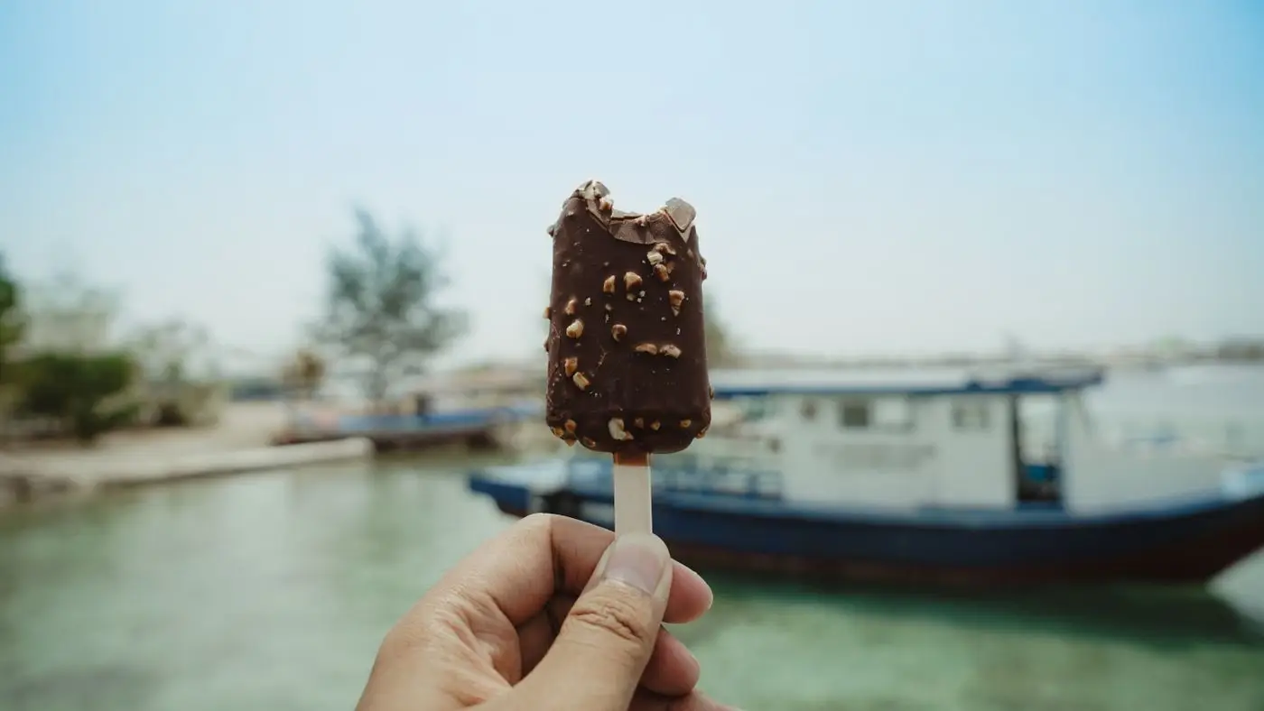 Hand holding a bitten chocolate-coated ice cream bar on a stick by the water