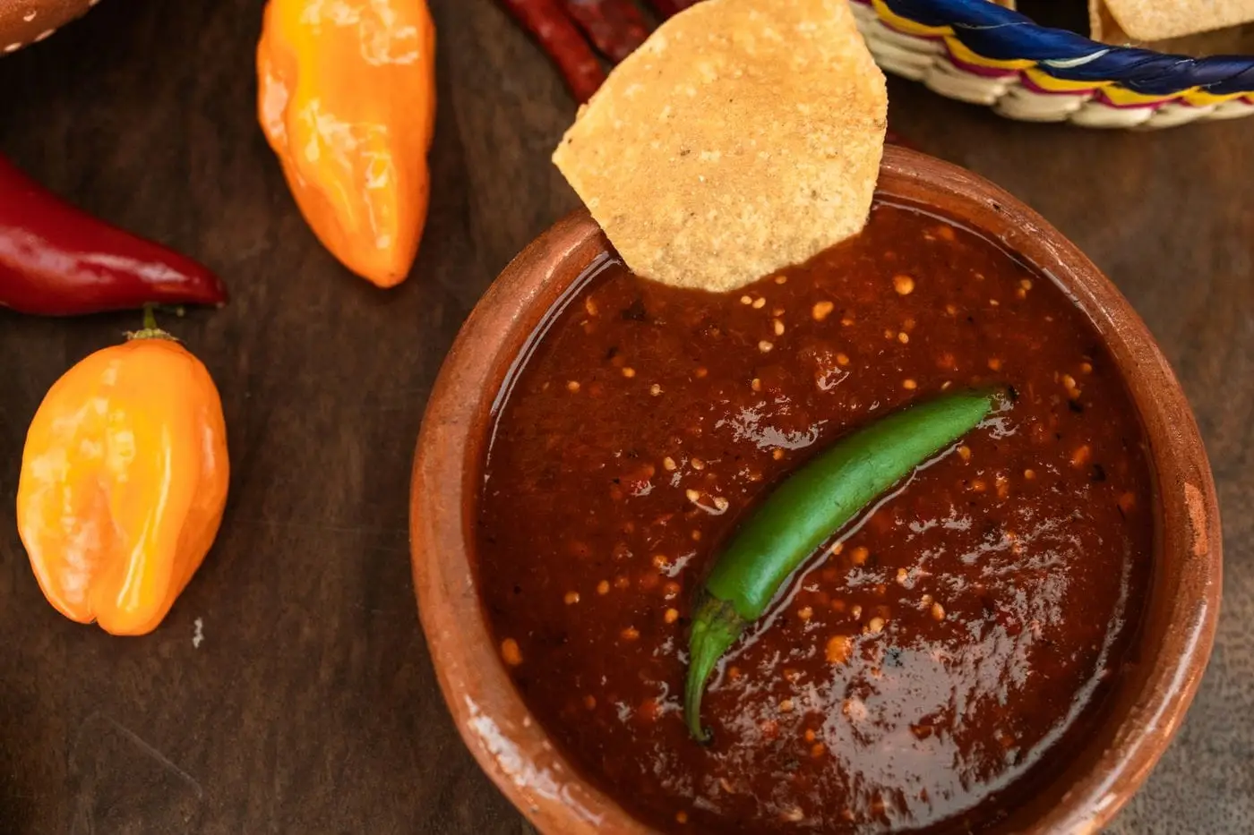 Bowl of red salsa with a green chili and tortilla chip beside peppers