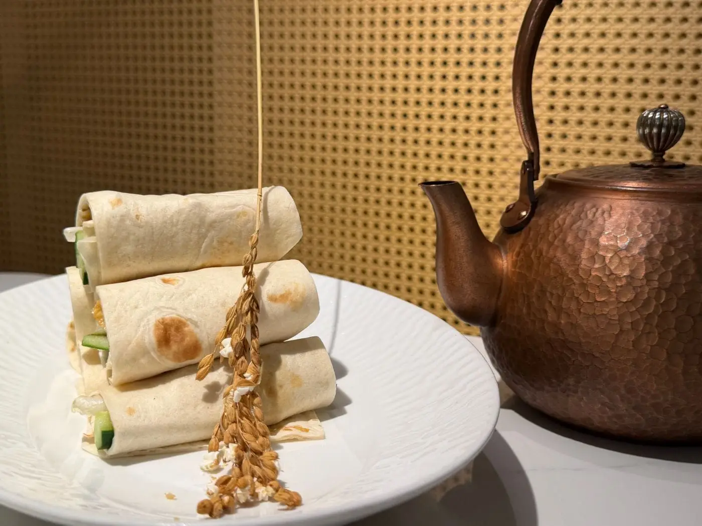 Stacked rolled flour tortilla wraps on a white plate beside a copper teapot